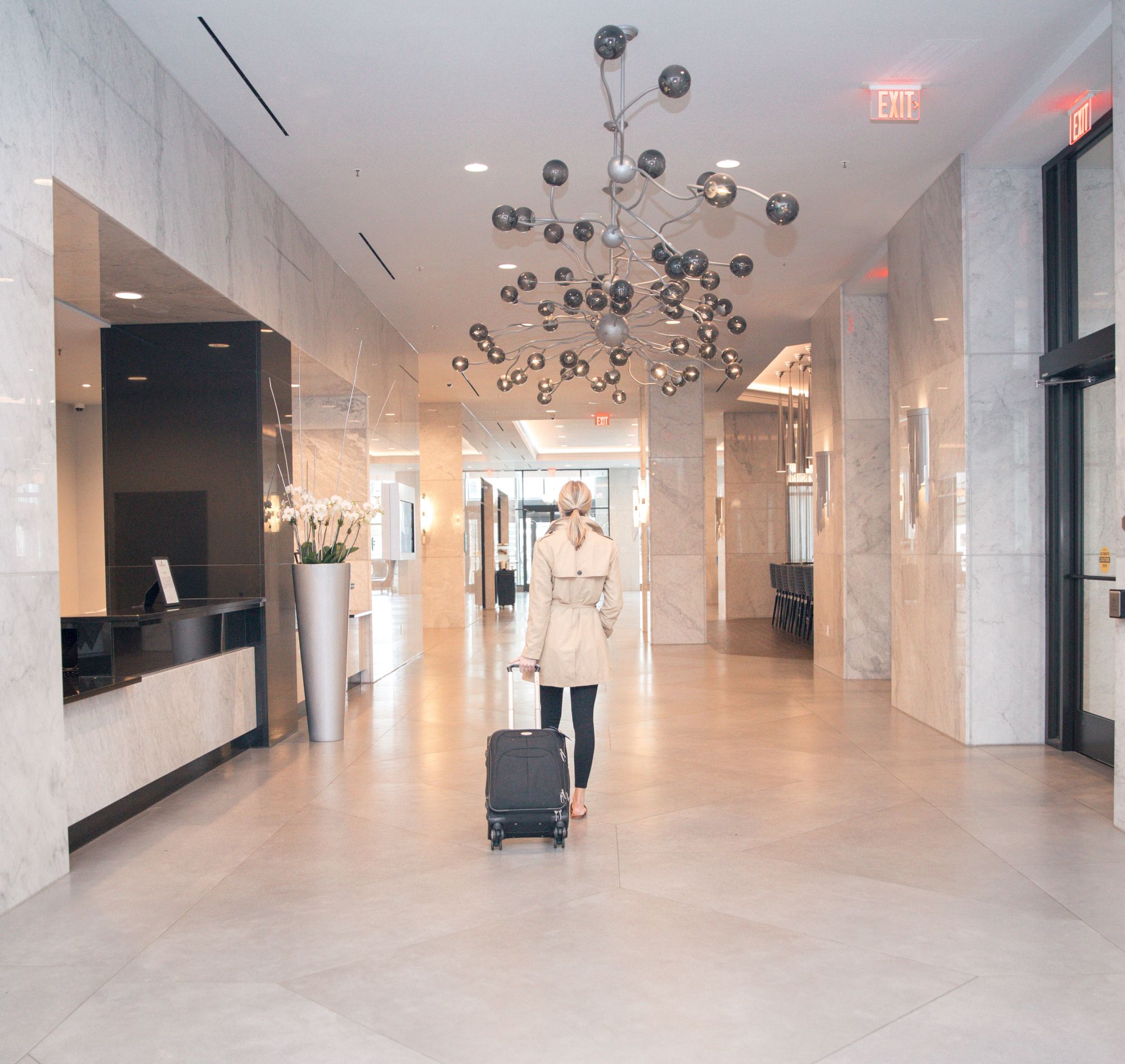 A person in a beige coat walks with a suitcase through a modern, marble-floored hallway with a decorative chandelier.