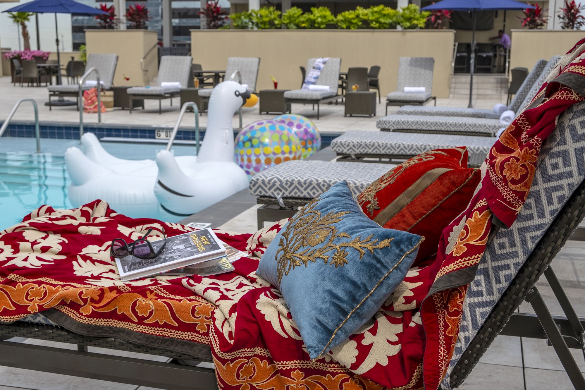 A poolside scene with lounge chairs, vibrant towels, pillows, a large swan float, sunglasses, and a magazine on a chair by the poolside.