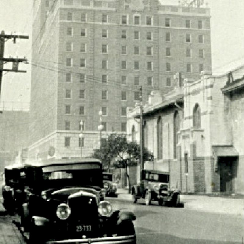 A black-and-white image shows vintage cars parked on a street with a tall building and power lines in the background.
