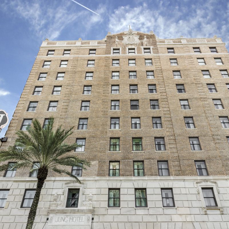 The image shows a tall, historic building with multiple windows, a palm tree, and a sign that reads “JUNG” against a partly cloudy sky.