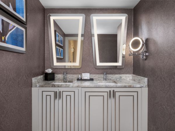 The image shows a bathroom with two lit mirrors above a double sink vanity, decorated with artwork and a textured wall.