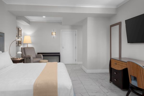 The image shows a modern hotel room with a bed, armchair, lamp, TV, desk, and chair on a tiled floor.