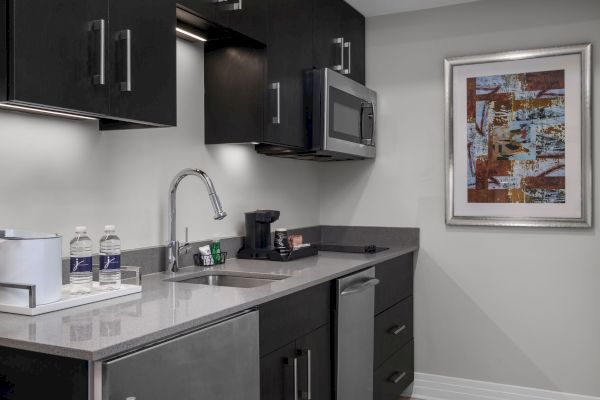 A modern kitchenette with a sink, microwave, coffee maker, two water bottles, and abstract art on the wall.