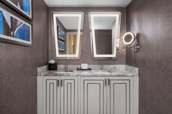 This image shows a modern bathroom vanity with dual sinks, two illuminated mirrors, a magnifying mirror, and decorative wall art.