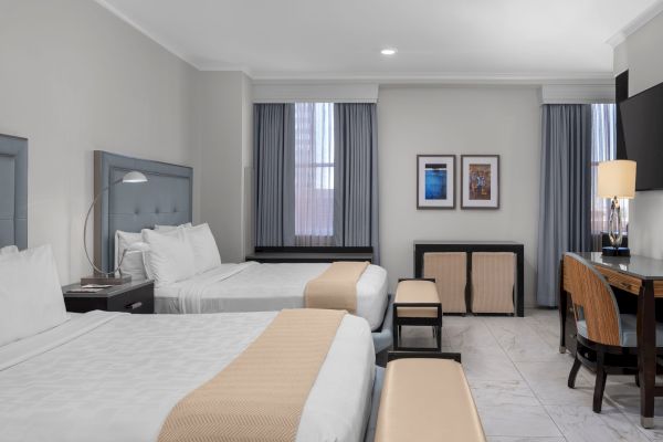 The image shows a modern hotel room with two beds, a desk, and two chairs, featuring neutral colors and framed artwork on the wall.