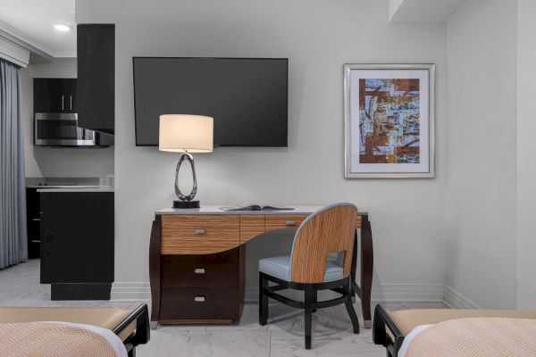 A hotel room with a desk, chair, lamp, and TV. There's abstract art on the wall. Kitchen area with microwave visible.