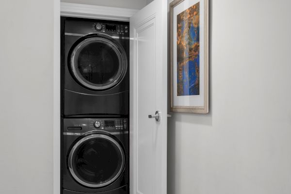 A stacked washer and dryer set with a white door beside a colorful framed picture on the wall.