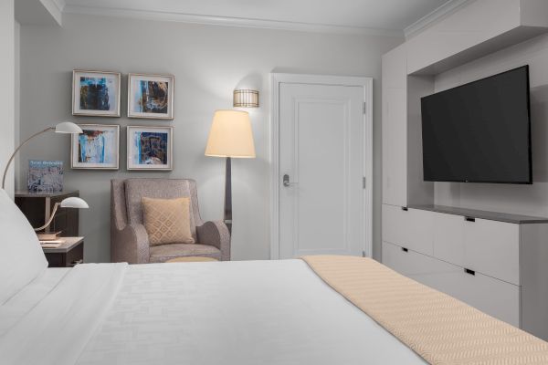 A cozy room featuring a bed, armchair, four wall artworks, a floor lamp, and a TV. A "New Orleans" book sits on the side table.