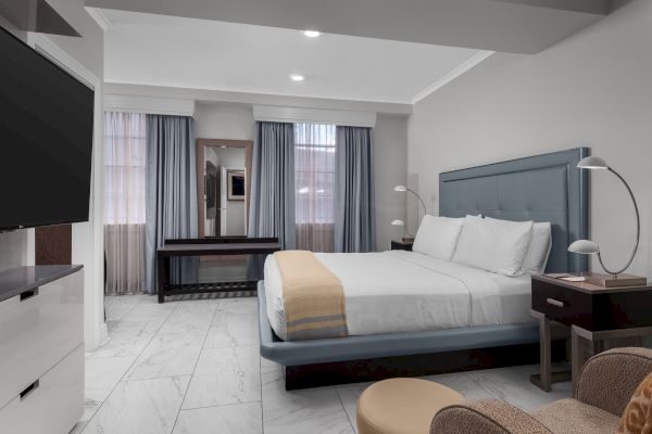 A modern bedroom with a large bed, side tables, lamps, a mirror, blue curtains, and a wall-mounted TV, featuring marble floors.