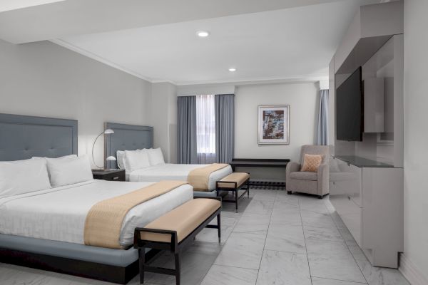 A modern hotel room with two beds, a TV, armchair, benches, and a window with curtains.