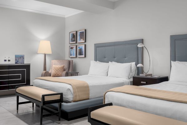 The image shows a hotel room with two beds, a chair, a lamp, and framed pictures on the wall.