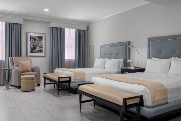 The image shows a hotel room with two double beds, a chair, an ottoman, a side table, and a framed artwork on the wall.