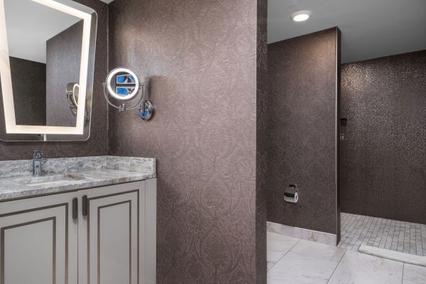 The image shows a modern bathroom with a marble countertop, lit mirror, wall-mounted magnifying mirror, and a walk-in shower.