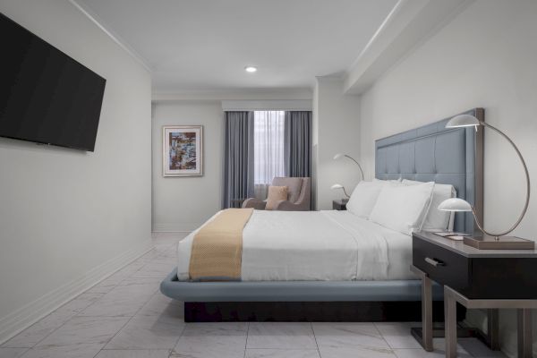 The image shows a modern bedroom with a bed, bedside lamps, an armchair, a framed picture, and a wall-mounted TV.