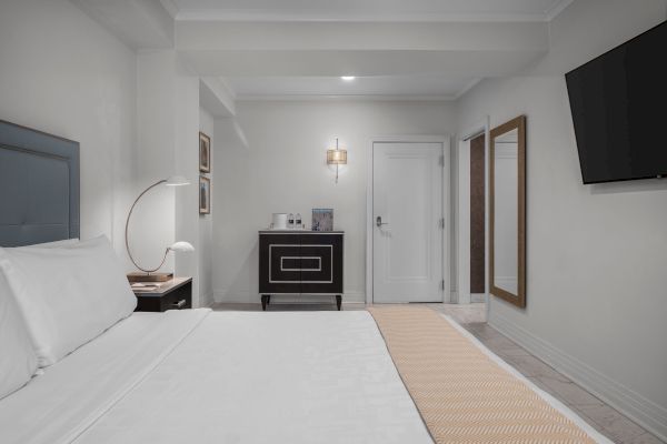 A modern hotel room with a neatly made bed, a TV on the wall, a side table, a cabinet, and a minimalist decor.