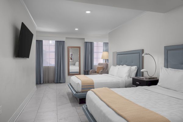 The image shows a hotel room with two beds, a wall-mounted TV, a mirror, armchair, side table, and large windows with curtains.