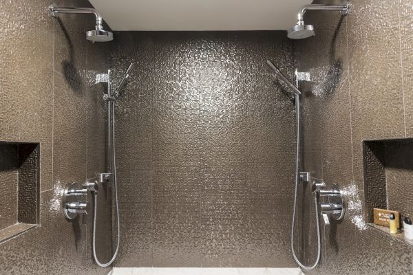The image shows a modern shower space with two showerheads and handheld sprays, set against a textured wall with built-in shelves.
