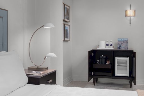 A modern hotel room with a bed, bedside lamp, framed art, minibar with snacks, drinks, and a brochure on top.