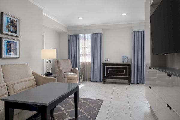 A modern living room with gray curtains, beige sofas, a black coffee table, framed artwork, and a flat-screen TV on the wall.