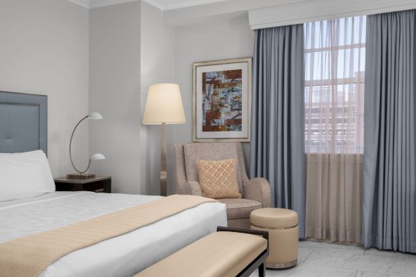 A cozy hotel room with a bed, armchair, lamp, decorative cushion, artwork, and window with blue curtains.