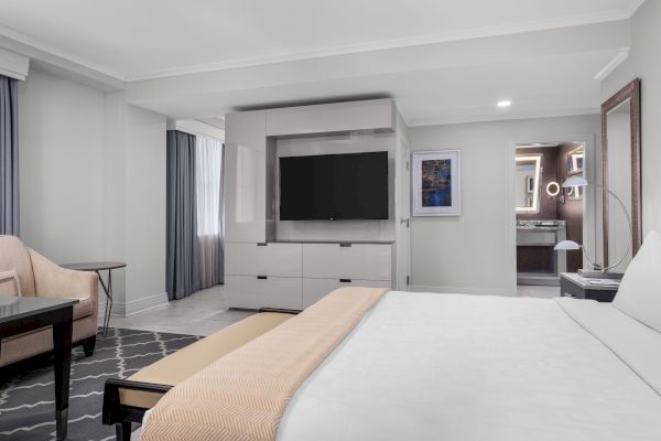 A modern hotel room with a bed, TV, table, armchair, and bathroom with a lighted mirror in view.