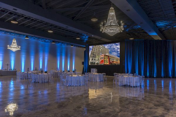 A spacious event hall with chandeliers, elegant tables, blue lighting, and a large screen displaying a city scene with a red streetcar.