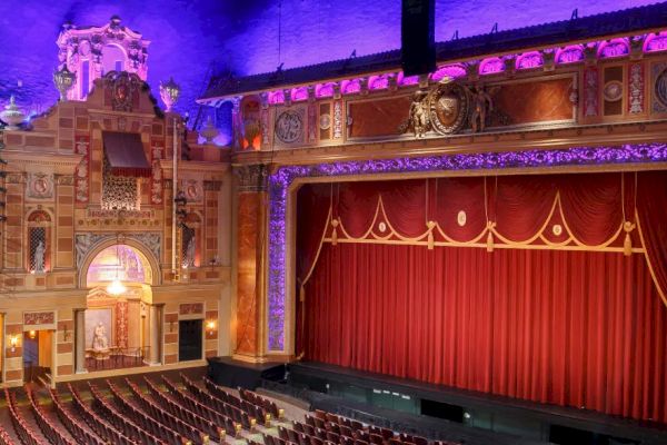 The image shows an ornate theater with a red curtain, intricate architecture, and tiered seating under a purple-lit ceiling.