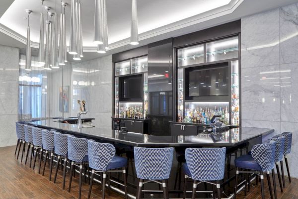 A modern bar with a large countertop, high-backed blue chairs, two TVs, and an array of hanging lights.