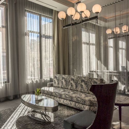 An elegant living room with a patterned sofa, modern chairs, a glass table, large windows, and stylish spherical chandeliers.
