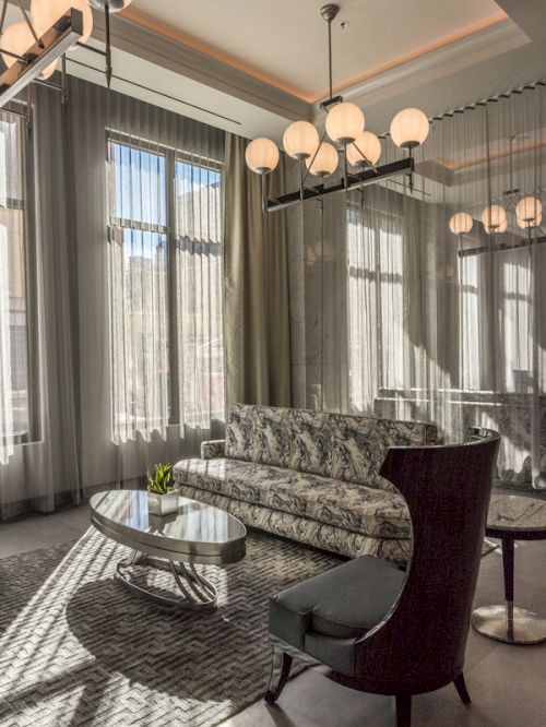 An elegant living room with a patterned sofa, modern chairs, a glass table, large windows, and stylish spherical chandeliers.