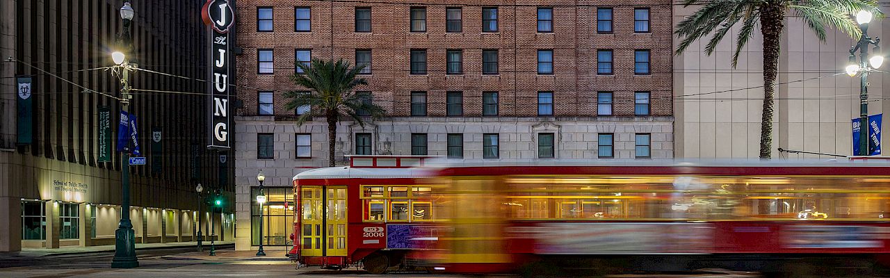 A historic brick building with palm trees in front and a streetcar moving past, under a cloudy evening sky.