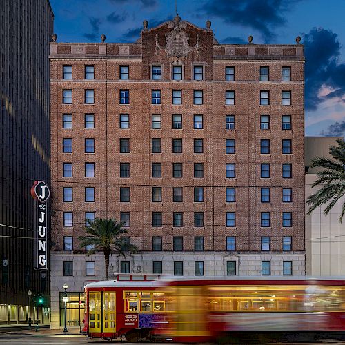 A historic brick building with palm trees in front and a streetcar moving past, under a cloudy evening sky.