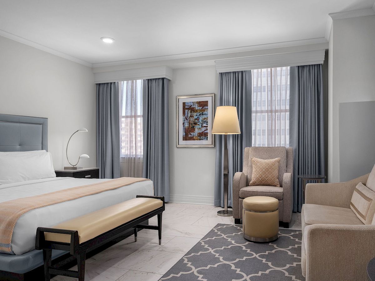 A modern hotel room with a bed, armchair, sofa, and a small ottoman. It includes a floor lamp, nightstand, and framed art.