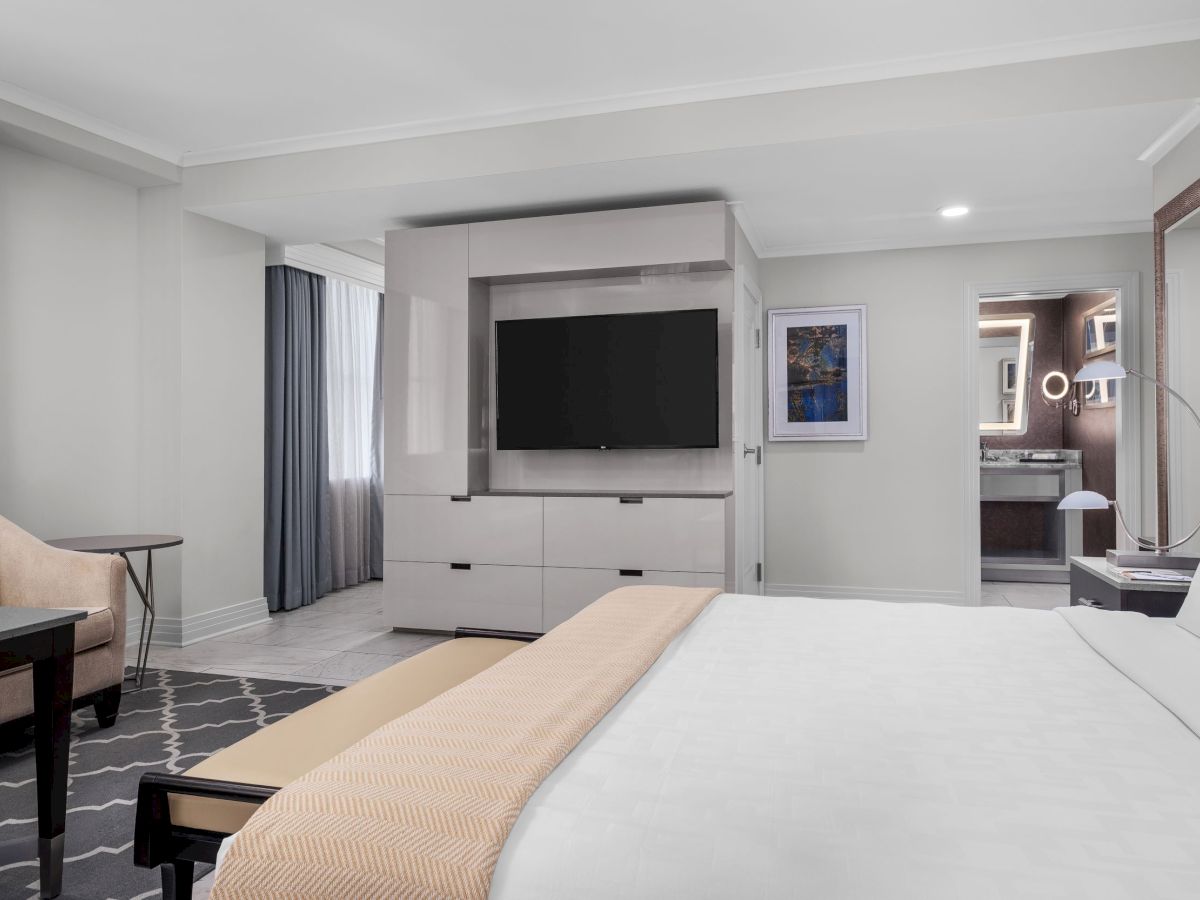 The image shows a modern hotel room with a bed, TV, armchair, and a bathroom with stylish lighting fixtures.