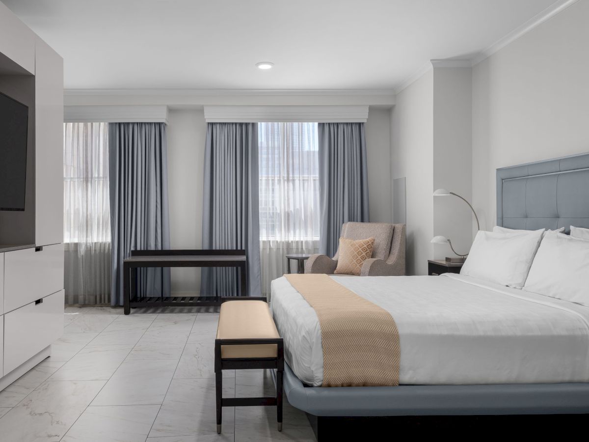 A modern hotel room with a bed, armchair, TV, and large windows with curtains, featuring a sleek, minimalist design.