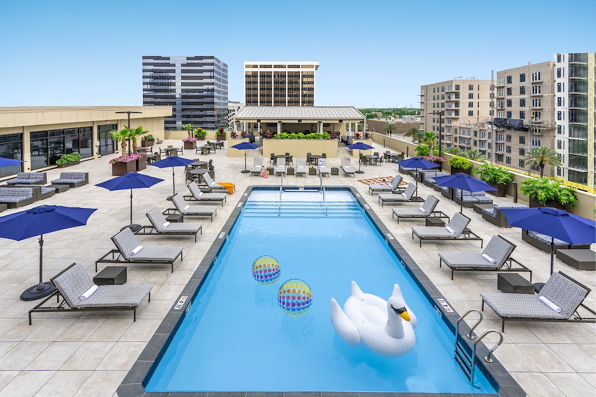 Rooftop pool with a large swan float, lounge chairs, and blue umbrellas. Surrounded by urban buildings under a clear blue sky.