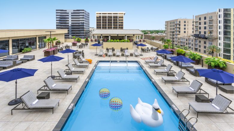 Rooftop pool with a large swan float, lounge chairs, and blue umbrellas. Surrounded by urban buildings under a clear blue sky.