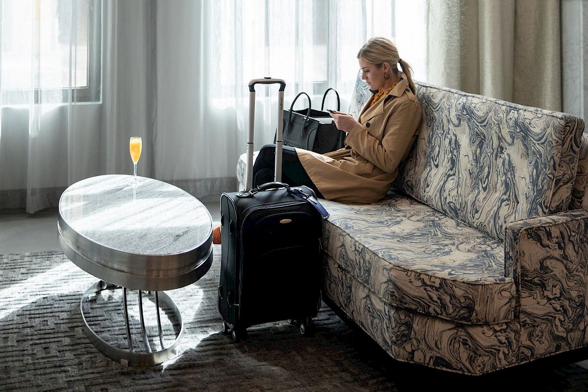 A person sits on a couch, looking at a phone. There's a suitcase nearby and a drink on the table in a well-lit room.
