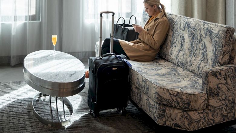A person sits on a couch, looking at a phone. There's a suitcase nearby and a drink on the table in a well-lit room.