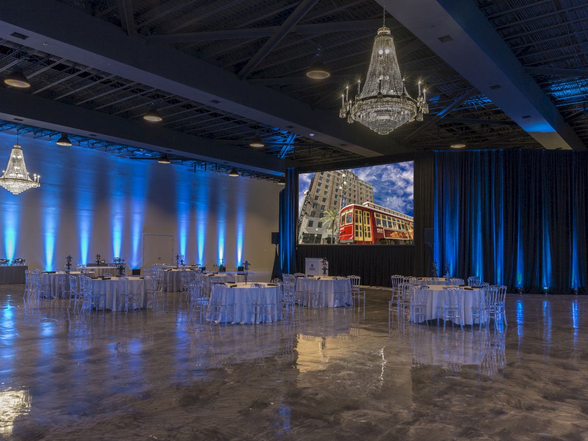 An elegant event hall with chandeliers, round tables, blue lighting, and a large screen displaying a streetcar.