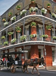A brick building with ornate iron balconies covered in plants, and a horse-drawn carriage passing by on the street.