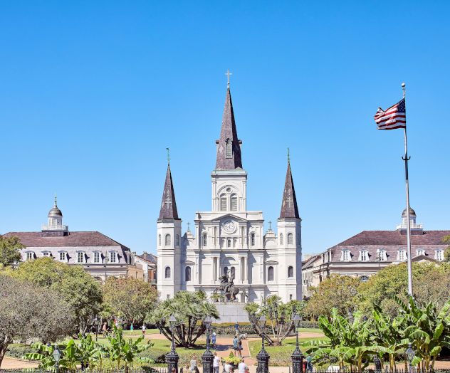 The image shows St. Louis Cathedral in New Orleans with a clear blue sky and an American flag flying in front.