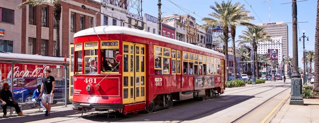 A red streetcar travels down a city street lined with palm trees and buildings, with people visible nearby at a tram stop.