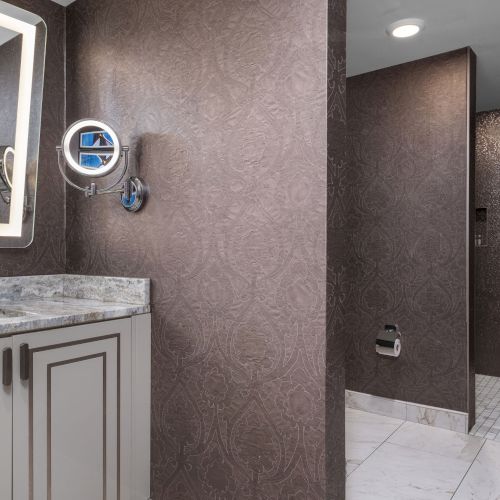 This image features a bathroom with a countertop, a lighted mirror, brown walls, tiled floor, and a walk-in shower area.