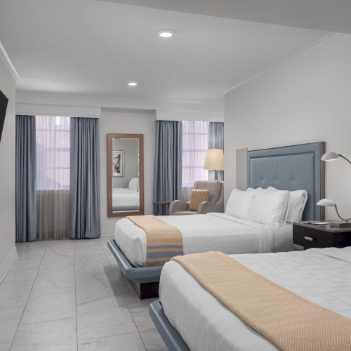 The image shows a hotel room with two beds, a wall-mounted TV, a standing mirror, and a window with sheer curtains.