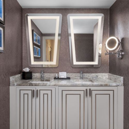 The image shows a bathroom with two sinks, tilted mirrors, artwork, and a makeup light on the wall, with a patterned wallpaper finish.
