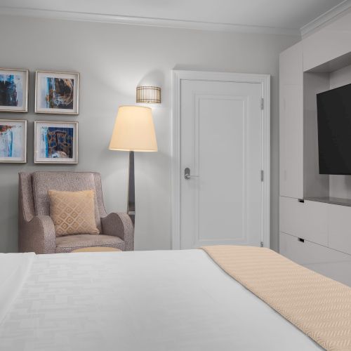 A modern hotel room with a bed, art on the wall, a floor lamp, an armchair, and a large flat-screen TV on a cabinet.