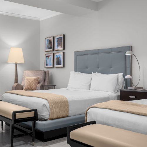 A hotel room with two beds, nightstand, a dresser, and a lamp by an armchair; artwork decorates the wall.