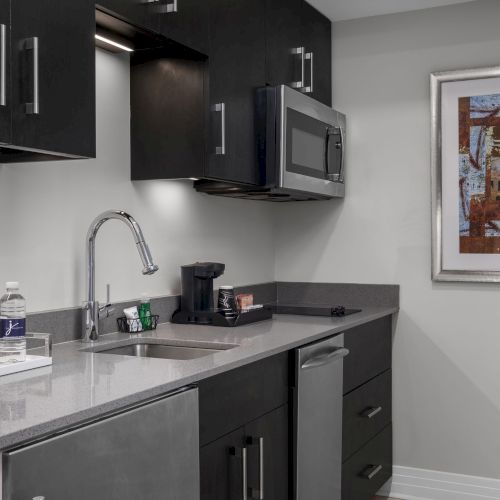 A modern kitchenette with dark cabinets, a sink, microwave, coffee maker, water bottles, and framed artwork on the wall.