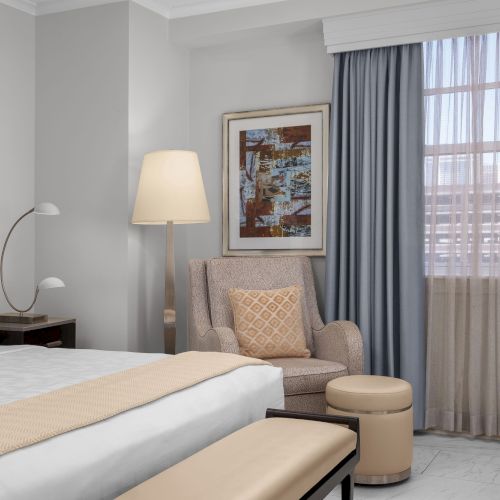 A hotel room with a bed, armchair, lamp, artwork, and window curtains. The decor is in neutral tones, creating a relaxing atmosphere.